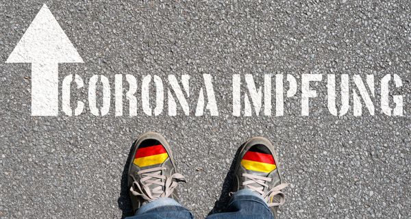 Schriftzug Corona-Impfung auf den Boden aufgesprüht mit zwei Schuhen davor.