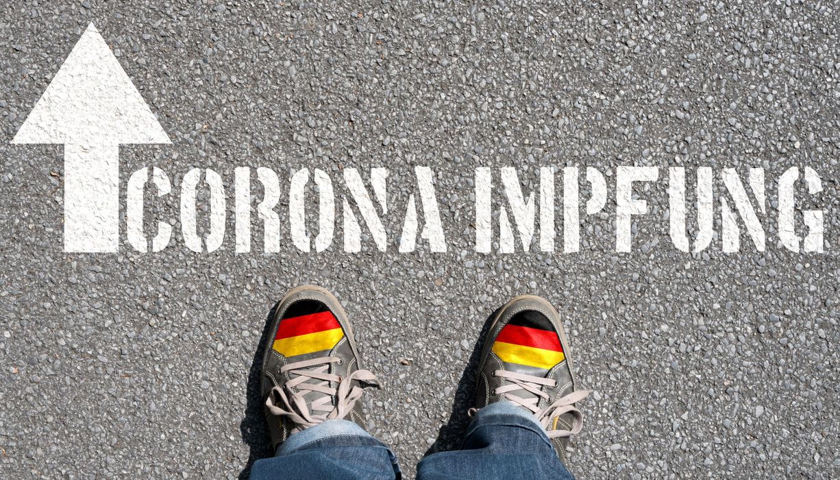 Schriftzug Corona-Impfung auf den Boden aufgesprüht mit zwei Schuhen davor.