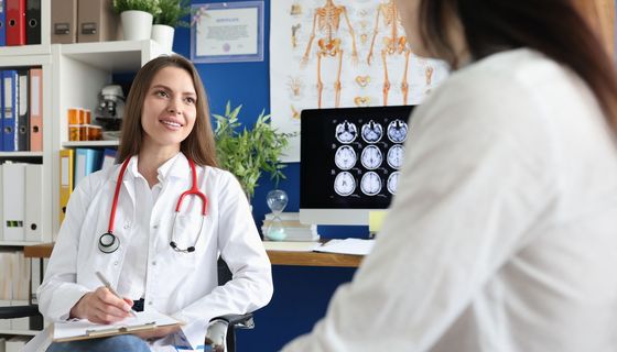 Eine Frau sitzt bei einer Ärztin, die eine Anamnese durchführt. Im Hintergrund sind Gehirnscans sichtbar.