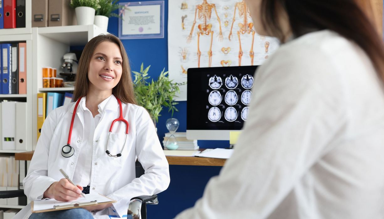 Eine Frau sitzt bei einer Ärztin, die eine Anamnese durchführt. Im Hintergrund sind Gehirnscans sichtbar.