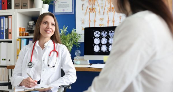 Eine Frau sitzt bei einer Ärztin, die eine Anamnese durchführt. Im Hintergrund sind Gehirnscans sichtbar.