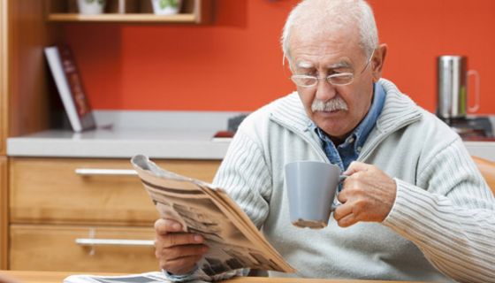Senior am Küchentisch beim Zeitunglesen, in der rechten Hand die Zeitung, in der linken einen Kaffeehumpen