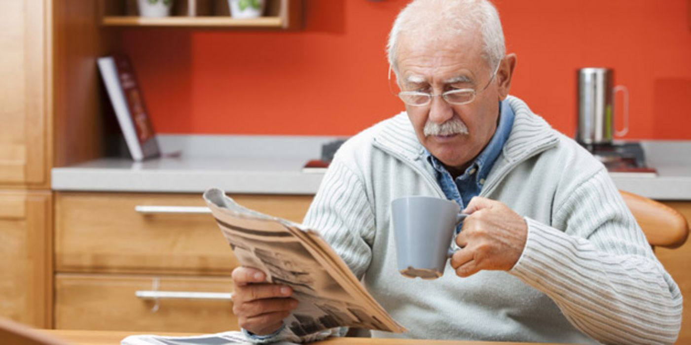 Senior am Küchentisch beim Zeitunglesen, in der rechten Hand die Zeitung, in der linken einen Kaffeehumpen
