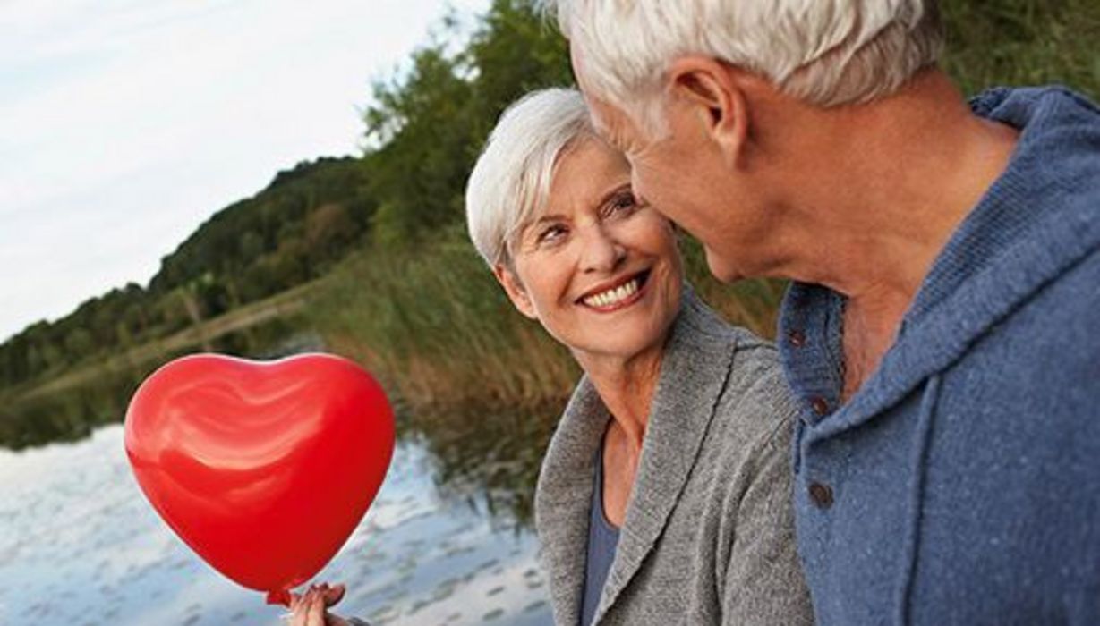 Attraktive grauhaarige Frau lacht grauhaarigen Mann an, der von schräg hinten zu sehen ist, Hintergrund Wasser und Wald; Frau hält roten Herzluftballon in einer Hand