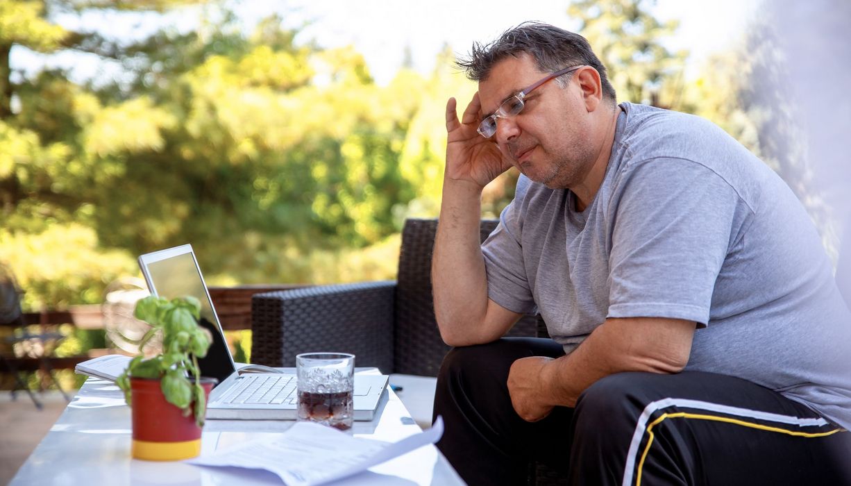 Mann mit Übergewicht, sitzt draußen auf der Terrasse vor seinem Laptop.
