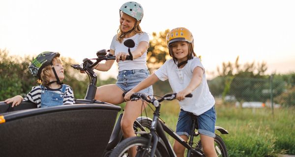 Junge Mutter mit Lastenrad und einem Kind darin, daneben ein weiteres Kind auf dem Fahrrad.