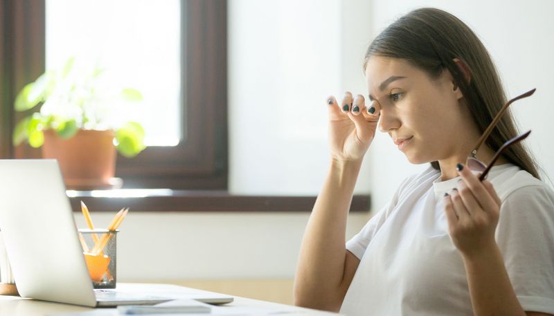 Frau vor dem Laptop reibt sich das Auge, in der anderen Hand hält sie die Brille.