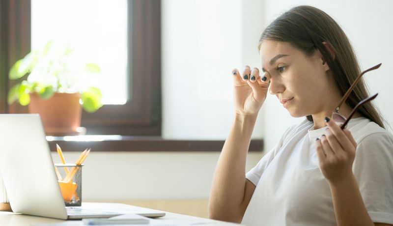 Frau vor dem Laptop reibt sich das Auge, in der anderen Hand hält sie die Brille.