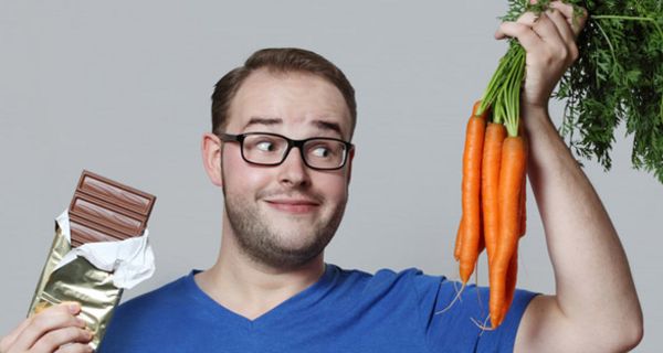 Junger Mann mit blauem T-Shirt hat in der einen Hand ein Bund Möhren, in der anderen eine Tafel Schokolade