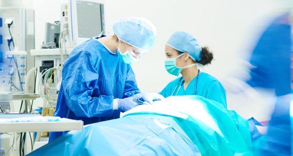 Patient liegt abgedeckt im Operationsaal, eine Chirurgin arbeitet am Patientin. Eine Op-Schwester überprüft die Werte.