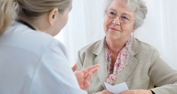 Ältere weißhaarige Dame freundlich lächelnd mit Rezept in der Hand im Beratungsespräch bei Ärztin (rechter Bildrand von hinten seitlich zu sehen)