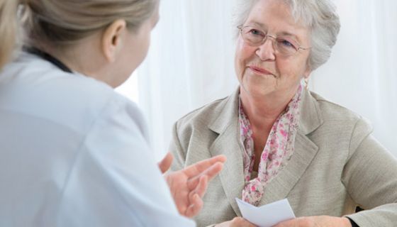 Ältere weißhaarige Dame freundlich lächelnd mit Rezept in der Hand im Beratungsespräch bei Ärztin (rechter Bildrand von hinten seitlich zu sehen)