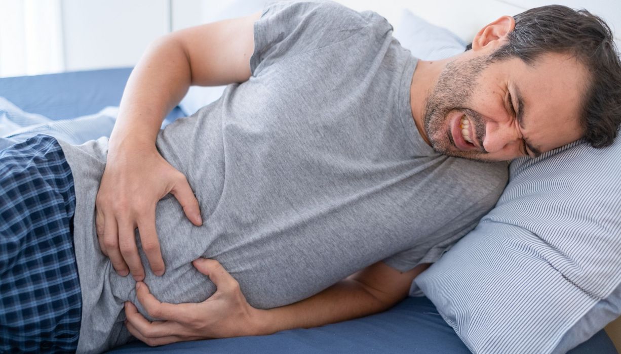 Mann krümmt sich vor Schmerzen auf dem Bett und hält sich den Bauch.