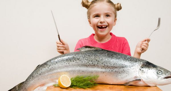 Blondes Mädchen lachend mit Messer und Gabel, auf dem Tisch vor sich einen großen Lachs