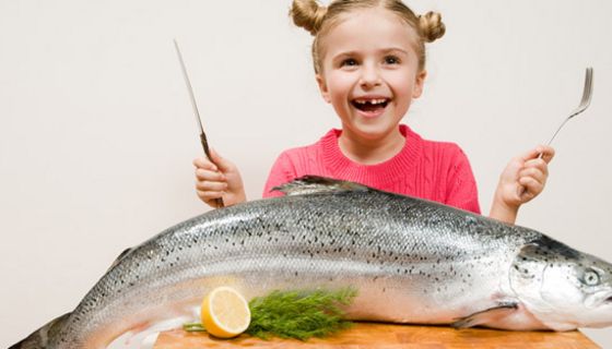 Blondes Mädchen lachend mit Messer und Gabel, auf dem Tisch vor sich einen großen Lachs