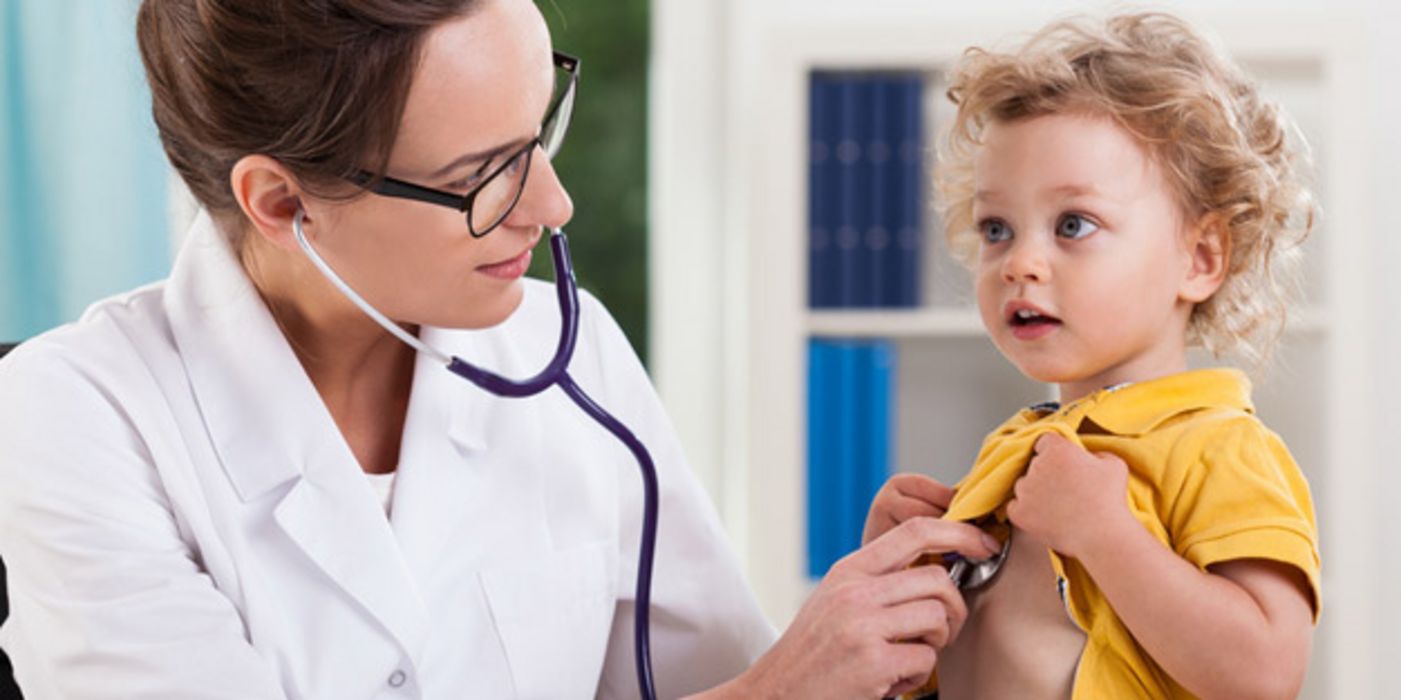 Junge Ärztin horcht mit Stethoskop Brust von blondgelocktem Kind (ca. 3 bis 4) ab. Gelbes Poloshirt wird vom Kind dazu hochgehalten