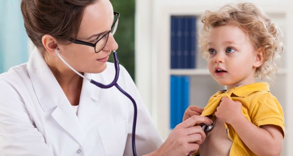Junge Ärztin horcht mit Stethoskop Brust von blondgelocktem Kind (ca. 3 bis 4) ab. Gelbes Poloshirt wird vom Kind dazu hochgehalten