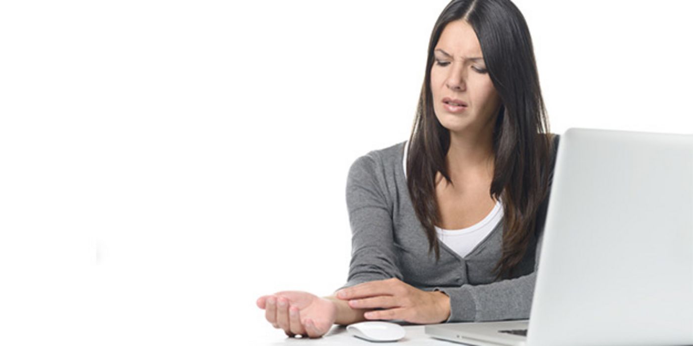 Dunkelhaarige Frau um die 30, weißer Laptop, weißer Schreibtisch, Maus, hält sich schmerzendes, rechtes Handgelenk