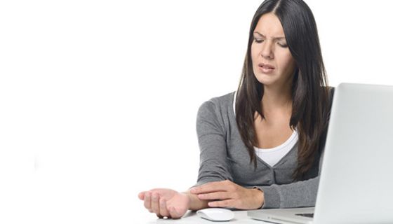 Dunkelhaarige Frau um die 30, weißer Laptop, weißer Schreibtisch, Maus, hält sich schmerzendes, rechtes Handgelenk