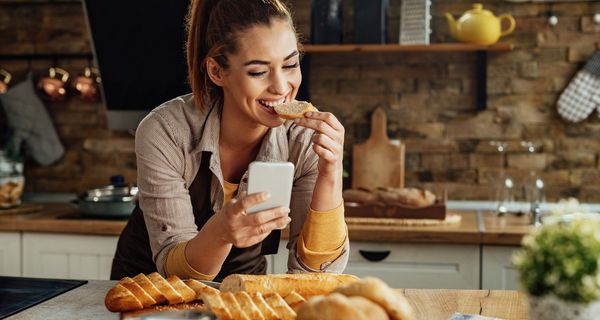 Frau, isst ein Baguette und schaut ins Handy.