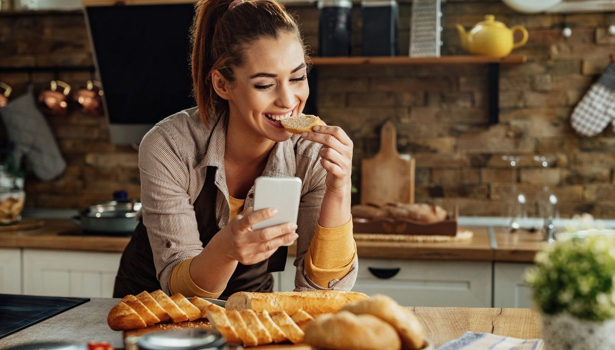 Frau, isst ein Baguette und schaut ins Handy.