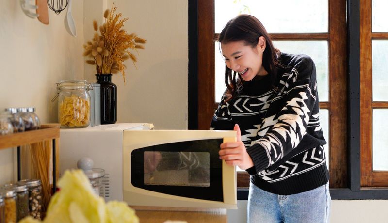 Junges Mädchen steht in einer Küche vor einer geöffneten Mikrowelle und lächelt.