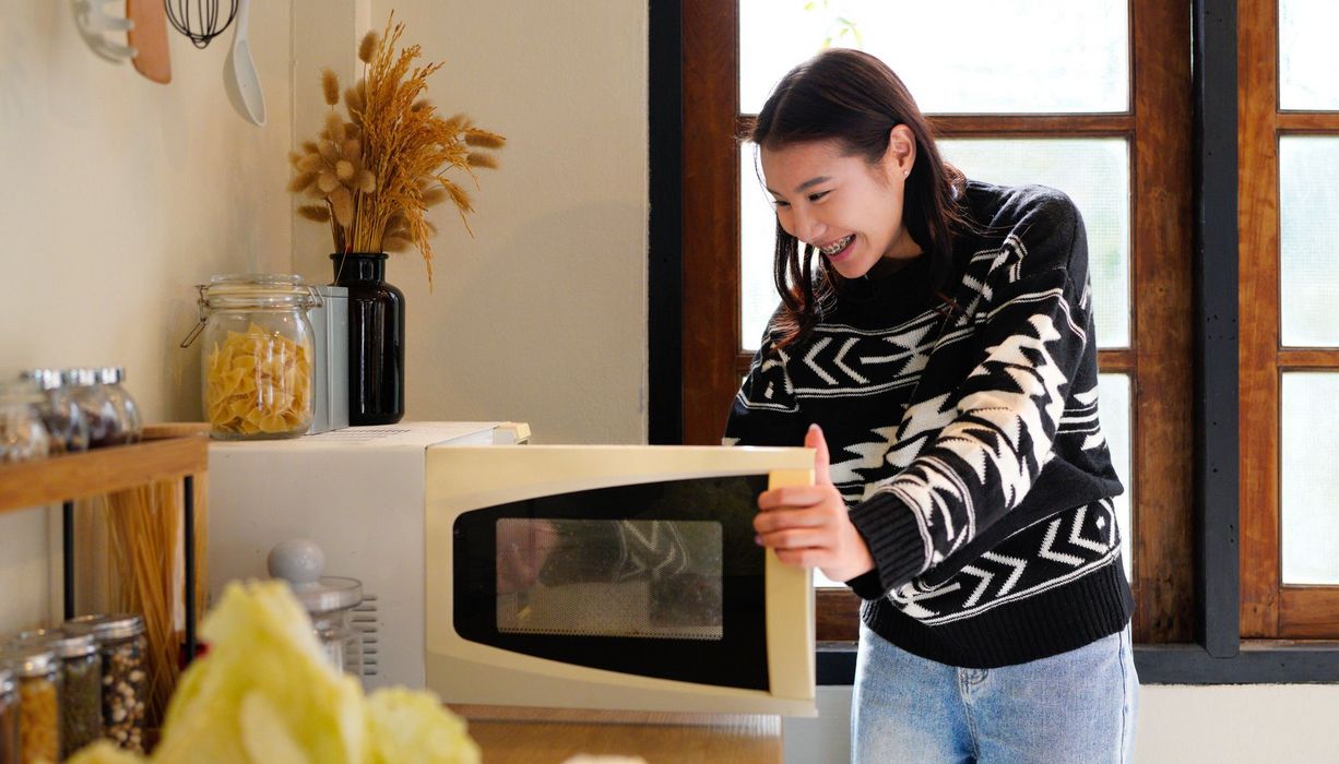 Junges Mädchen steht in einer Küche vor einer geöffneten Mikrowelle und lächelt.
