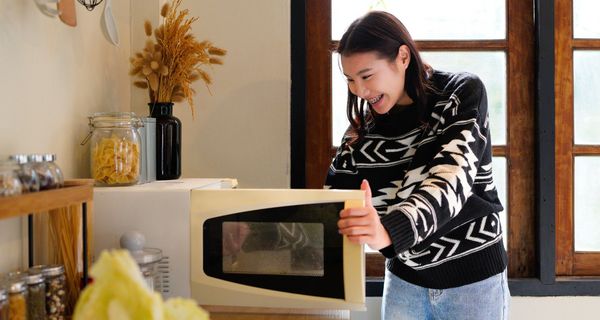 Junges Mädchen steht in einer Küche vor einer geöffneten Mikrowelle und lächelt.