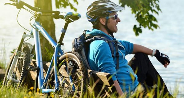 Mann um die 50 in Fahrradkleidung mit Helm lehnt an einem See am Baum, Mountainbike an die andere Seite des Baumes gelehnt. Abendstimmung