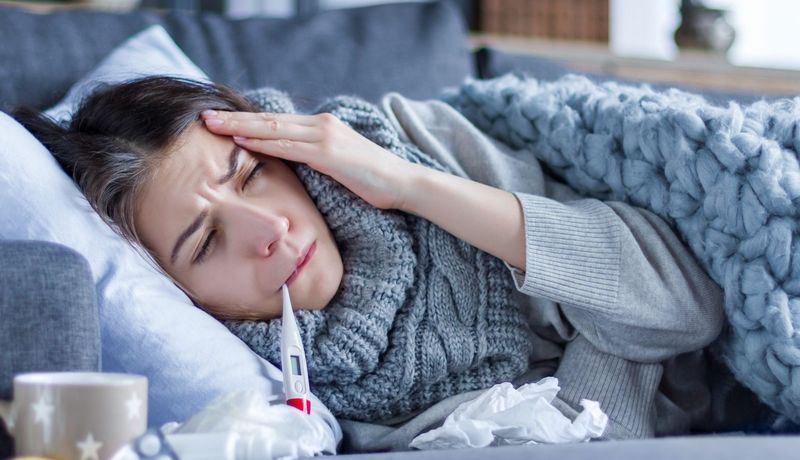 Blasse Frau liegt mit zusammengekniffenen Augen im Bett, eine Hand an ihrer Stirn, ein Thermometer hängt aus ihrem Mundwinkel.