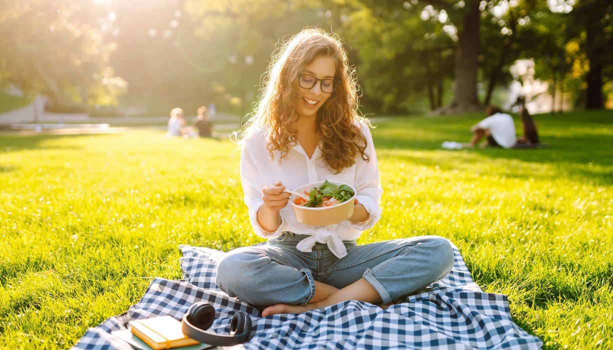 Eine fröhliche Frau mit Brille sitzt auf einer Matte in einem Park und isst an einem sonnigen Tag einen Salat.