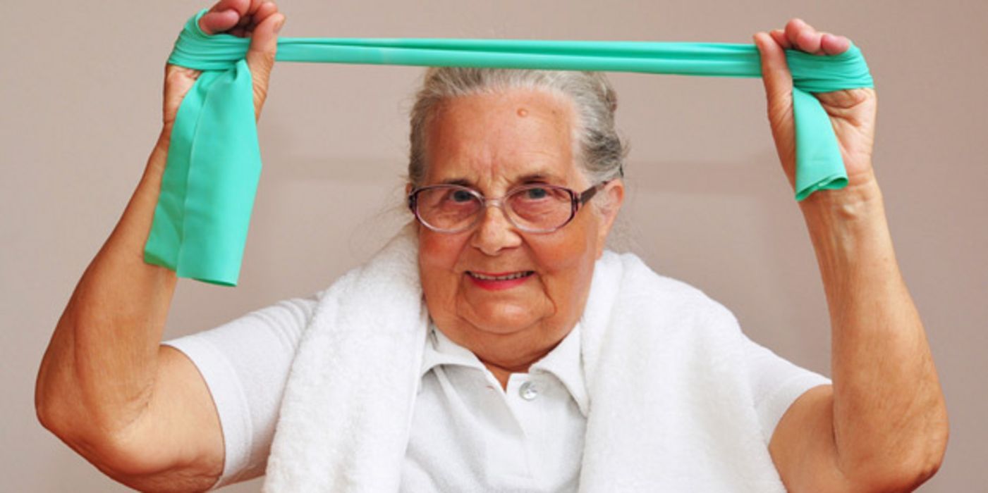 Frontalfoto einer grauhaarigen Seniorin, ca. Mitte 70, mit türkisfarbenem Terraband mit erhobenen Armen über dem Kopf dehnend
