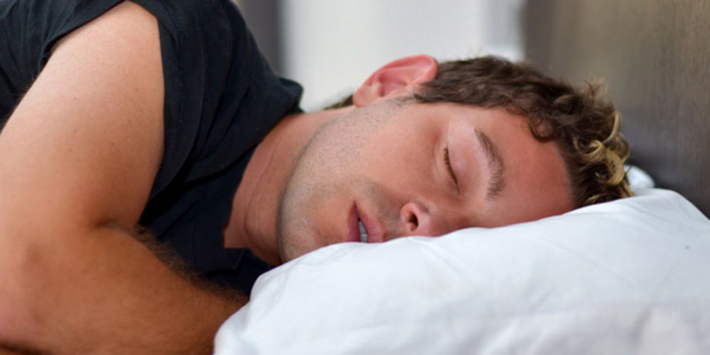 Nahaufnahme eines jungen, dunkelhaarigen Mannes (ca. Mitte/Ende 20), schwarzes Kurzarmshirt, schlafend auf der Seite liegend auf weißem Kopfkissen