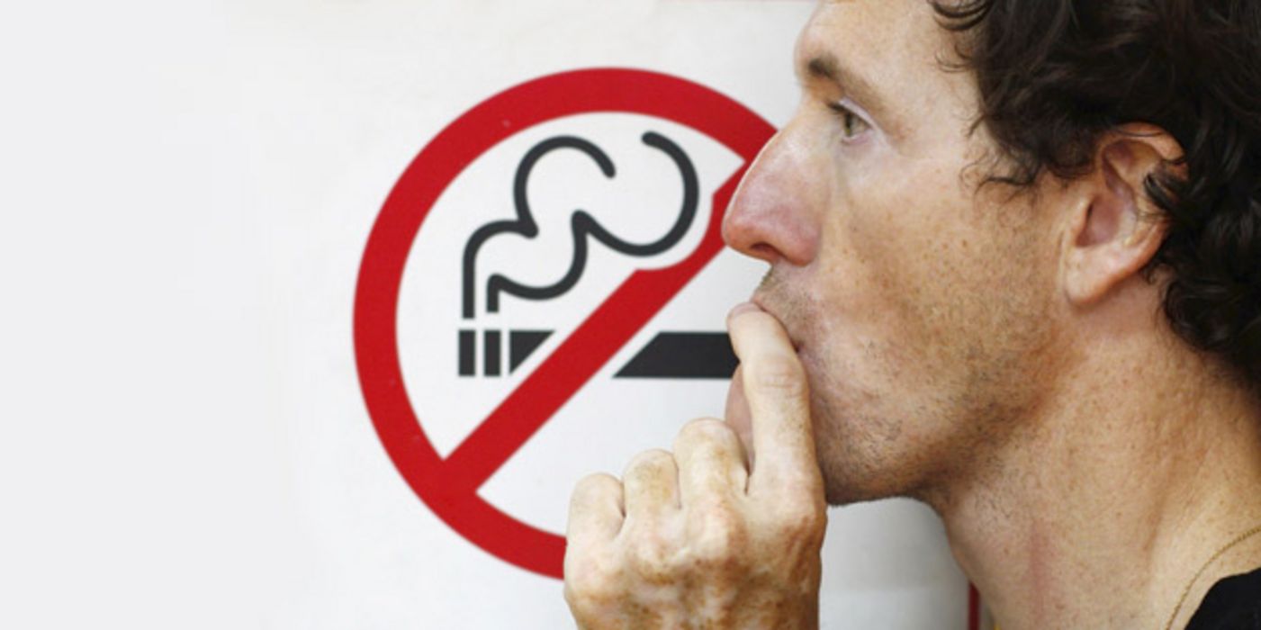 Mann mit Dreitagebart vor einem Rauchverbotsschild tut so als ob er rauchen würde
