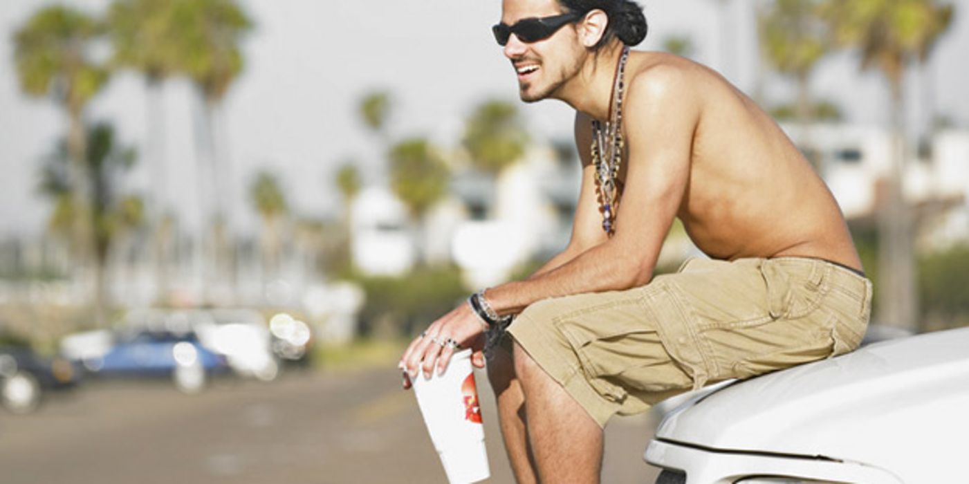 Junger Mann sitzt mit einem Softdrink in der Hand auf der Motorhaube eines Autos