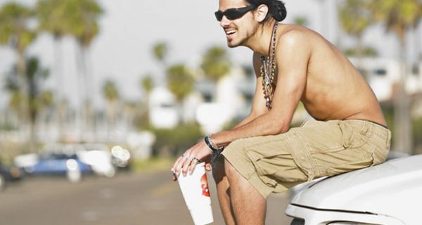 Junger Mann sitzt mit einem Softdrink in der Hand auf der Motorhaube eines Autos