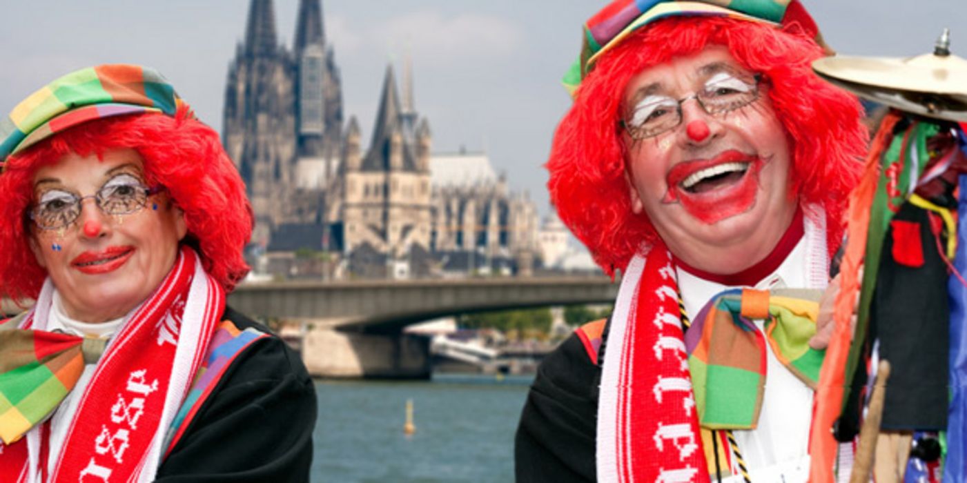 Karnevalistisches Paar in Clowsnsschminke und Hüten mit roten Perücken lackend vor Rhein und Kölner Dom