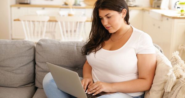 Übergewichtige Junge Frau, sitzt auf dem Sofa, Laptop auf dem Schoß.