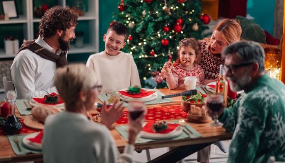 Familie rund um den geschmückten Esstisch beim Weihnachtsfest: Eine Frau füttert ein Kind, ein Junge spricht mit einem Erwachsenen, zwei ältere Personen unterhalten sich.