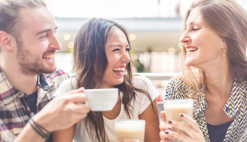 Zwei junge Frauen und ein junger mann trinken zusammen Kaffee und lachen.