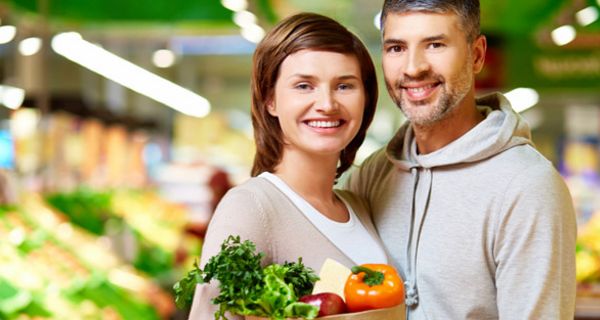 Junges lächelndes Paar vor Gemüseregal, Frau trägt eine Papiertüte mit Gemüse in der Hand