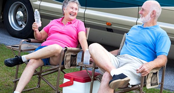 Lachendes Rentnerpaar auf Campingstühlen vor Auto mit Kühltasche