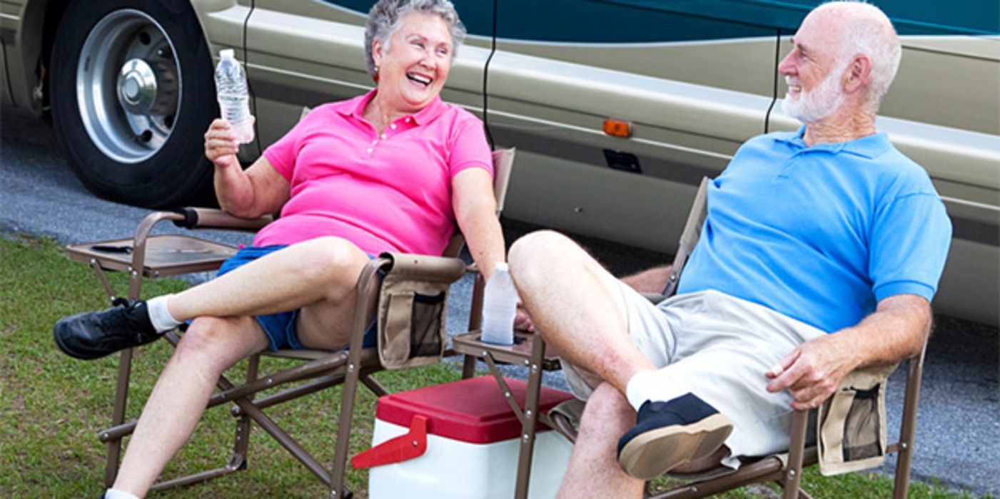Lachendes Rentnerpaar auf Campingstühlen vor Auto mit Kühltasche