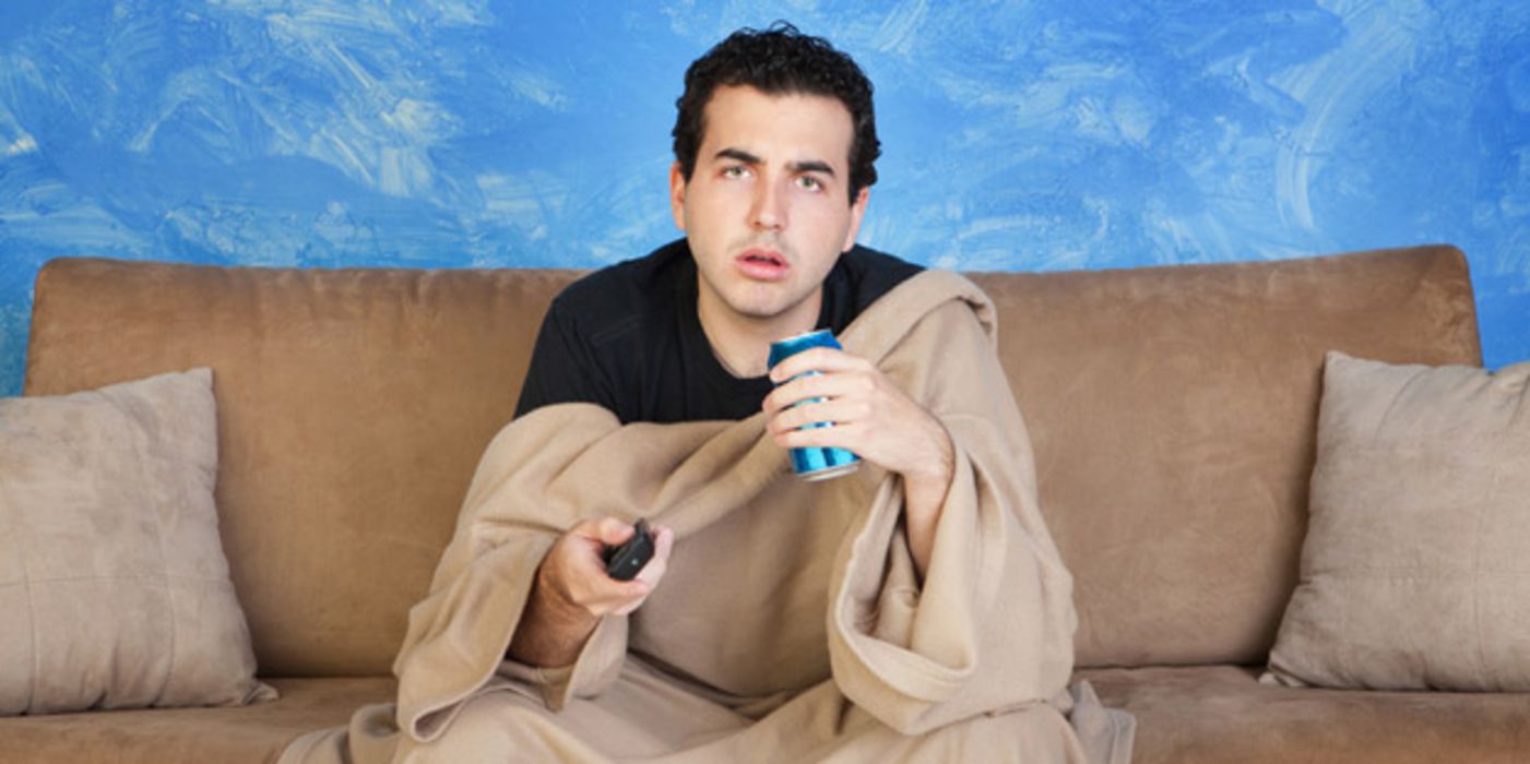 Jüngerer dunkelhaariger Mann sitzt mit verpeiltem Gesichtsausdruck auf einer Couch, Fernbedienung in der einen, Getränkedose in der anderen Hand, über den Knien eine Decke