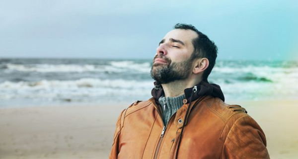 Mann am Meer (Oberkörper), rehbraune Lederjacke, schwarze Haare, Vollbart, ca. Ende 30, atmet mit geschlossenen Augen tief ein