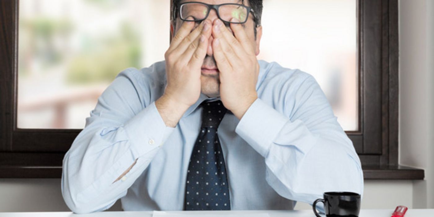 Frontalfoto: Mann an Schreibtisch, hellblaues Hemd, dunkelblau-weiß gemusterter Schlips, Ellenbogen auf Schreibtisch gestützt, reibt sich die Augen, Hornbrille auf Stirn geschoben