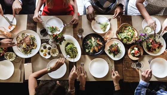 Blick auf einen reichgefüllten Tisch von oben, an dem sieben Personen sitzen. Von ihnen sind nur die Arme zu sehen. 