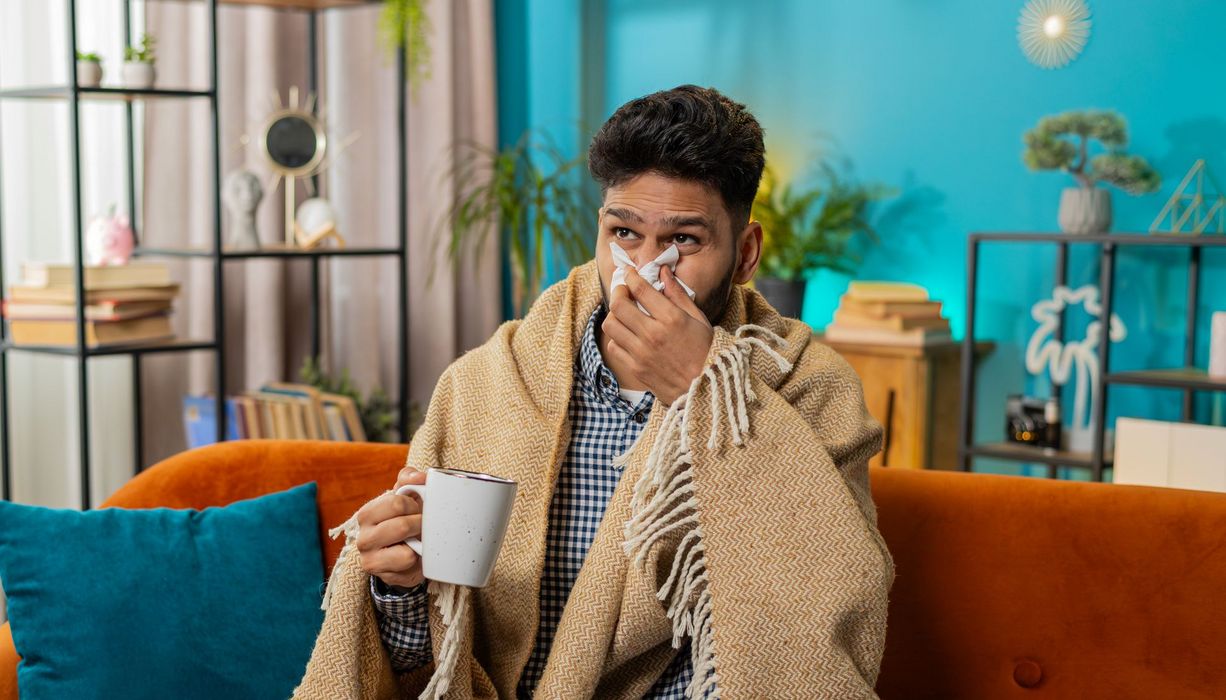 Ein kranker Mann sitzt auf einem Sofa und schnäuzt sich die Nase.