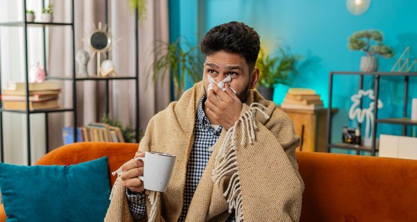 Ein kranker Mann sitzt auf einem Sofa und schnäuzt sich die Nase.