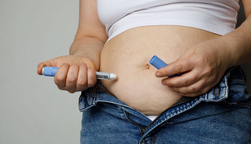 Eine übergewichtige Frau spritzt sich mit der Rechnet Hand ein Medikament in den Bauch. 